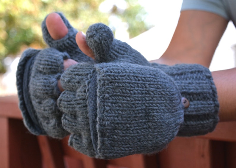 May include: Gray knitted convertible mittens with a button closure. The mittens have a flap that can be folded back to expose the fingers, transforming them into fingerless gloves. The knitted texture is visible, and the mittens appear warm and cozy.