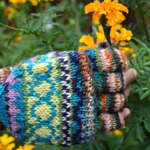 Könnte beinhalten: Bunte, gestrickte Fingerhandschuhe mit einem geometrischen Muster in Blau, Grün, Lila und Gelb. Die Handschuhe wirken warm und sind für kühleres Wetter geeignet. Im Hintergrund sind gelbe Blumen und grünes Laub zu sehen.