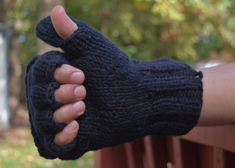 May include: A black knitted fingerless glove, with a thumb hole and a ribbed design. The glove is made of a dark, possibly wool, material. The person's hand is wearing the glove, with the thumb up.