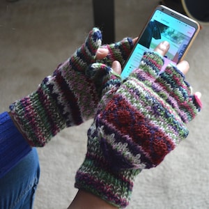 May include: Fingerless knitted gloves in a colorful striped pattern of red, green, purple, and white. The gloves are being worn while holding a smartphone. The cuffs are a darker shade of purple.