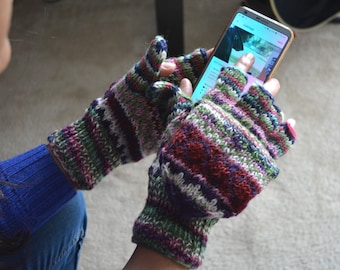 May include: Fingerless knitted gloves in a colorful striped pattern of red, green, purple, and white. The gloves are being worn while holding a smartphone. The cuffs are a darker shade of purple.