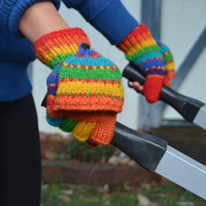 May include: Rainbow-striped knitted mittens with a convertible flap, held by a person wearing a blue sweater. The mittens feature a button closure on the flap and are made of colorful yarn in shades of red, orange, yellow, green, and blue.