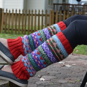 May include: A pair of colorful knitted leg warmers. The leg warmers have a red cuff and a patterned design in shades of blue, purple, green, and red. The person is wearing black leggings and gray shoes.