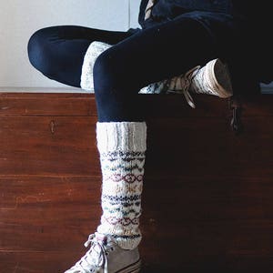 May include: A person wearing black pants and white knitted leg warmers with a colorful pattern. The person is sitting on a wooden chest with a metal latch.
