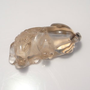 May include: A translucent, light brown, carved quartz figurine of a reclining lion. The figurine is detailed with intricate carvings and has a smooth, polished finish.