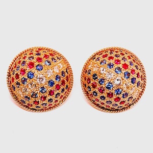 May include: A pair of gold earrings with a round, dome-shaped design. The earrings are set with a variety of colorful gemstones, including red rubies, blue sapphires, and white diamonds.