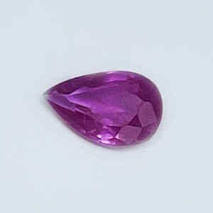 May include: A pear-shaped gemstone with a deep purple hue. The gemstone is faceted and has a high polish.