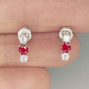 May include: A pair of silver drop earrings, each featuring a vertical arrangement of three stones: a clear round stone at the top, a vibrant red round stone in the center, and a clear round stone at the bottom, secured with a post backing.