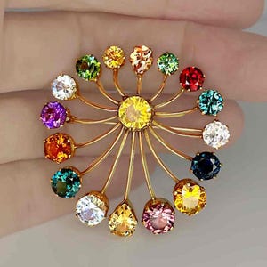May include: A gold-toned brooch featuring a central yellow gemstone surrounded by a radial design of smaller, colorful gemstones. The brooch showcases a variety of colors, including red, green, blue, and purple, in a circular arrangement.