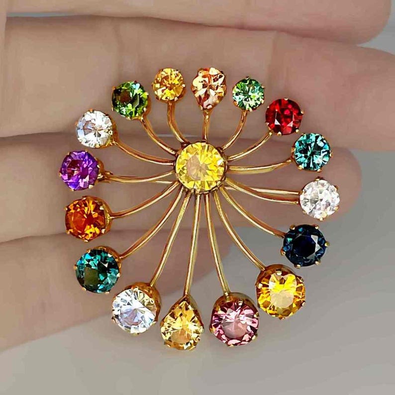 May include: A gold-toned brooch featuring a central yellow gemstone surrounded by a radial design of smaller, colorful gemstones. The brooch showcases a variety of colors, including red, green, blue, and purple, in a circular arrangement.