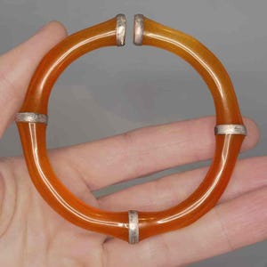 May include: Orange-amber colored bangle bracelet with silver-tone metal accents. The bracelet has a smooth, glossy finish and a curved, open design. The silver accents are at the joints of the bracelet.