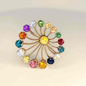 May include: A gold-toned brooch featuring a central yellow gemstone surrounded by a halo of colorful gemstones. The gemstones are in various colors, including purple, orange, green, and blue. The brooch has a floral or starburst design.