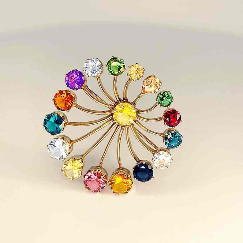 May include: A gold-toned brooch featuring a central yellow gemstone surrounded by a halo of colorful gemstones. The gemstones are in various colors, including purple, orange, green, and blue. The brooch has a floral or starburst design.