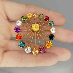 May include: A gold-toned brooch with a central yellow gemstone, surrounded by radiating gold wires. Each wire supports a different colored gemstone, including green, red, purple, and orange. The brooch is held in a hand against a gray background.