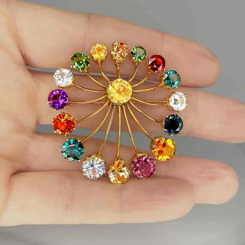 May include: A gold-toned brooch with a central yellow gemstone, surrounded by radiating gold wires. Each wire supports a different colored gemstone, including green, red, purple, and orange. The brooch is held in a hand against a gray background.