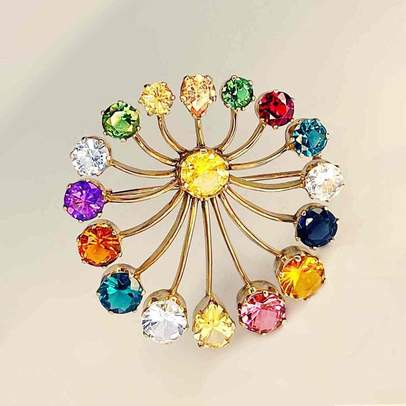 May include: A gold-toned brooch featuring a central yellow gemstone surrounded by a circular arrangement of colorful gemstones. The gemstones are in various colors, including green, red, blue, and purple. The brooch has a unique, radiant design.