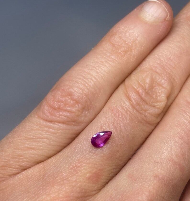 May include: A teardrop-shaped, deep pink gemstone with a faceted cut.