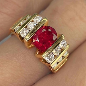 Può includere: Un anello d'oro con un grande rubino rosso circondato da diamanti più piccoli. L'anello ha un design unico con barre d'oro e una montatura a punta.