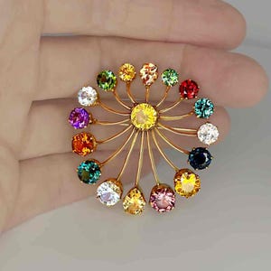 May include: A gold-toned brooch featuring a central yellow gemstone surrounded by a circular arrangement of colorful gemstones. The gemstones are in various colors, including red, green, blue, and purple. The brooch has a delicate, intricate design.