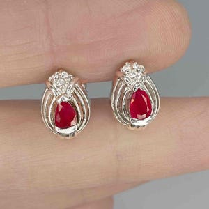 May include: A pair of silver earrings featuring a teardrop-shaped, vibrant red gemstone. The earrings have a detailed, openwork design with small, sparkling accents at the top. The gemstones are the focal point, set in a decorative silver setting.