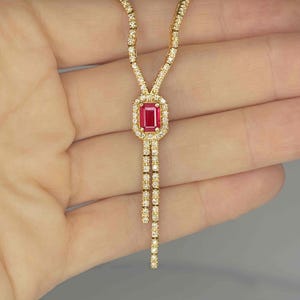 Puede incluir: Collar dorado con un colgante central rectangular de rubí, enmarcado por un halo de pequeñas gemas transparentes. La cadena está adornada con gemas transparentes, y el diseño incluye dos hilos colgantes.
