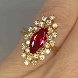 May include: A gold ring with a large, marquise-cut ruby and a halo of small, round diamonds. The ruby is a deep red hue, and the diamonds sparkle against the gold setting. The ring is on a finger.