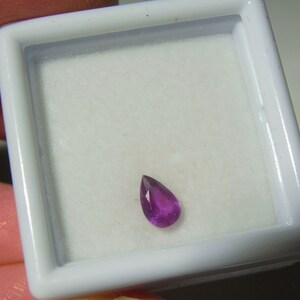 May include: A teardrop-shaped purple gemstone in a white jewelry box.
