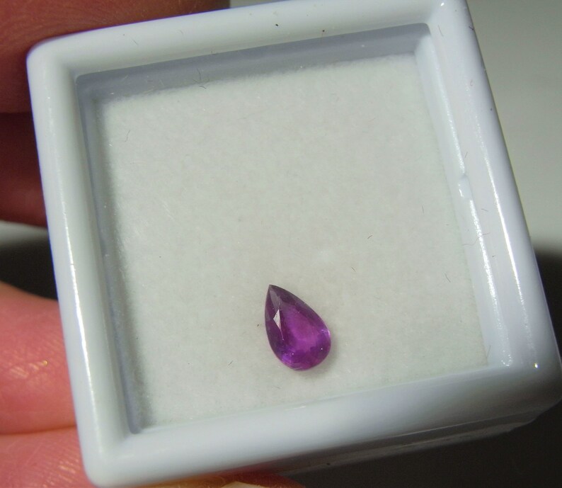 May include: A teardrop-shaped purple gemstone in a white jewelry box.