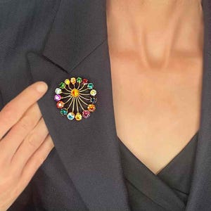 May include: A colorful brooch with a circular design. The brooch features a gold-tone metal frame with radiating lines and is adorned with a variety of colorful gemstones. The brooch is attached to a dark navy blazer.