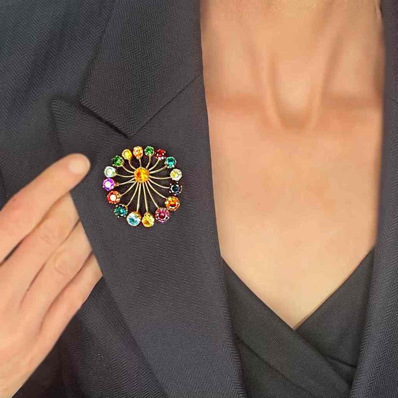May include: A colorful brooch with a circular design. The brooch features a gold-tone metal frame with radiating lines and is adorned with a variety of colorful gemstones. The brooch is attached to a dark navy blazer.