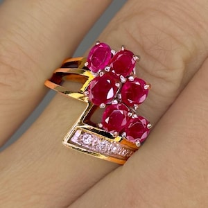 Op de afbeelding: Een gouden ring met een cluster van rode robijn edelstenen en een rij kleinere witte diamanten.
