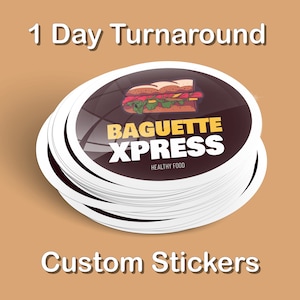 May include: A stack of round custom stickers with a white border and a black background. The sticker features a cartoon illustration of a baguette sandwich with the text "Baguette Xpress" in yellow and white letters. The text "Healthy Food" is printed in white below the logo.