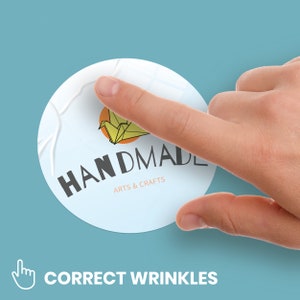 May include: A white round sticker with a yellow origami crane and the text "HANDMADE ARTS & CRAFTS". The sticker is slightly wrinkled and a finger is pressing down on it to smooth out the wrinkles.