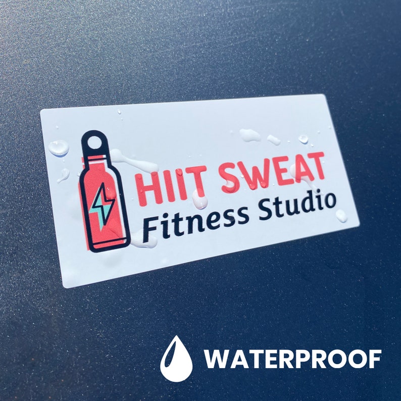May include: White sticker with red and black text that reads "HIIT SWEAT Fitness Studio". A red water bottle with a lightning bolt inside is on the left side of the sticker. The sticker is on a dark gray surface with a white water drop icon and the word "WATERPROOF" below the sticker.