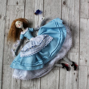 May include: A porcelain doll dressed in a blue and white dress with a white lace trim. The doll has red hair and is lying on a wooden surface.