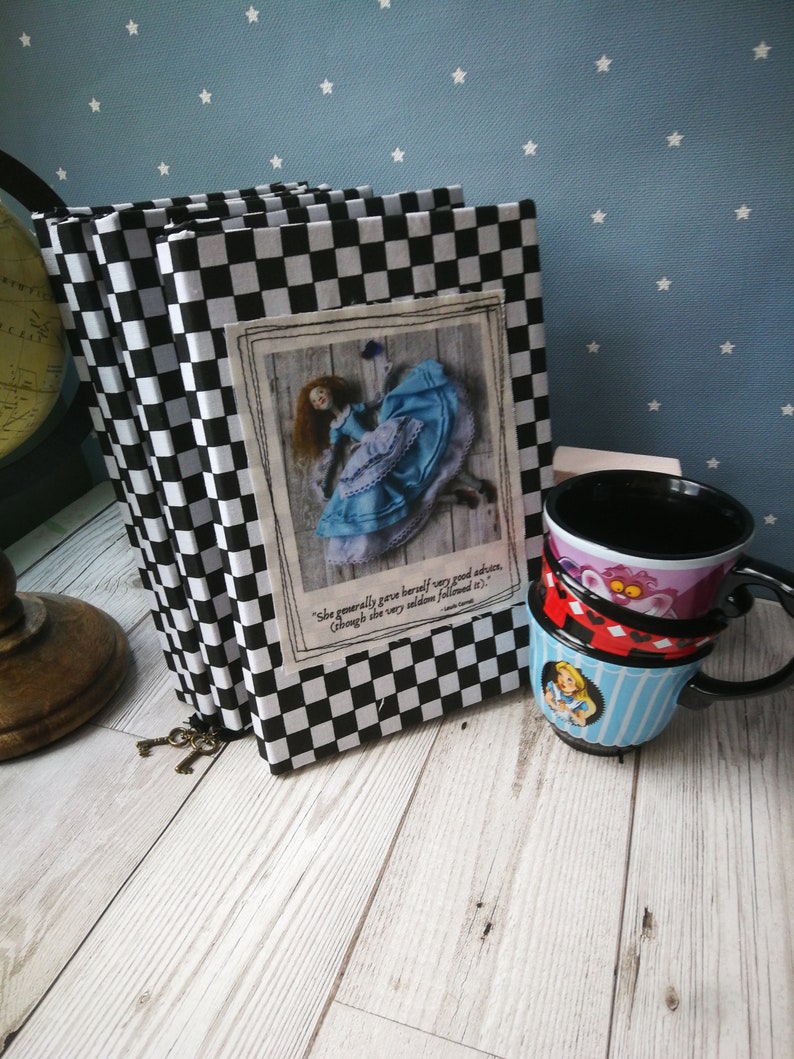May include: Three black and white checkered notebooks with a quote from Alice in Wonderland on the front cover. The quote reads "She generally gave herself very good advice, (though she very seldom followed it)." The notebooks are stacked on top of each other and have a key attached to the spine.