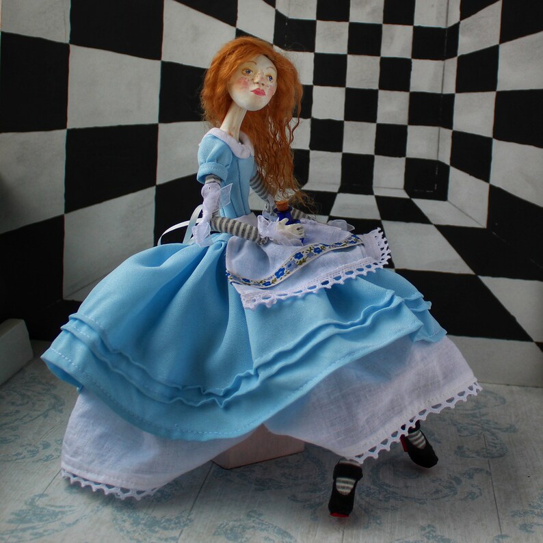 May include: A porcelain doll dressed as Alice in Wonderland, wearing a blue and white dress with a lace trim, sitting on a checkered floor.