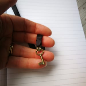May include: A gold-toned key charm on a black ribbon. The key has an ornate design and is a popular accessory for necklaces and bracelets.