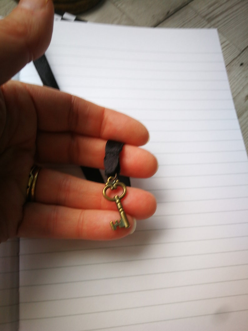 May include: A gold-toned key charm on a black ribbon. The key has an ornate design and is a popular accessory for necklaces and bracelets.