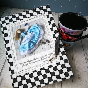 May include: A black and white checkered notebook with a quote from Lewis Carroll's Alice in Wonderland. The quote reads "She generally gave herself very good advice, (though she very seldom followed it)." The notebook features a picture of Alice falling down a rabbit hole.