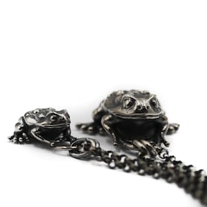 Könnte beinhalten: Eine Halskette aus dunkel silberfarbenem Metall mit zwei detaillierten Froschanhängern in unterschiedlichen Größen. Der kleinere Frosch ist an der Kette befestigt, während der größere Frosch als zentraler Anhänger dient. Die Kette besteht aus kleinen, runden Gliedern.