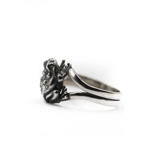 Puede incluir: Un anillo plateado con un diseño detallado de una rana. La rana está colocada en la parte superior del anillo, con los brazos extendidos. El anillo tiene una banda lisa y curvada. El fondo es blanco.