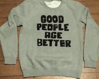 以下が含まれることがあります： 黒文字で「GOOD PEOPLE AGE BETTER」とプリントされた薄いグレーのスウェットシャツです。