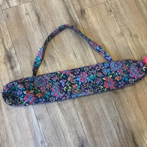 May include: A colorful yoga mat bag with a floral paisley pattern in shades of blue, purple, and red. The bag has a matching handle and a bright pink ribbon tie closure. The bag is designed to carry a yoga mat.