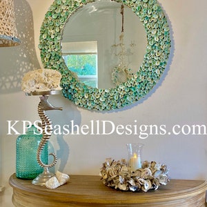 May include: A round mirror with a green and white seashell frame. The mirror is hanging on a wall above a wooden table with a seashell wreath and a silver seahorse statue.