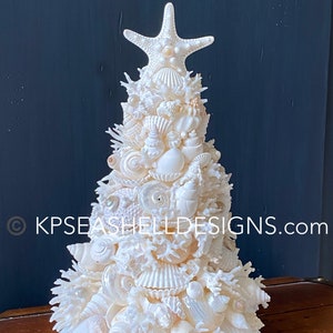 May include: A decorative Christmas tree made of various white seashells, coral, and a starfish topper. The tree is displayed on a wooden surface against a dark blue background. The tree is a unique coastal-themed decoration.