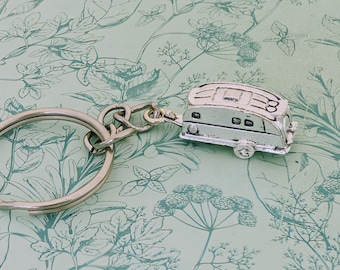 May include: Silver-colored camper keychain with a large key ring. The miniature camper has detailed windows and wheels. The keychain is on a light green background with a floral pattern.