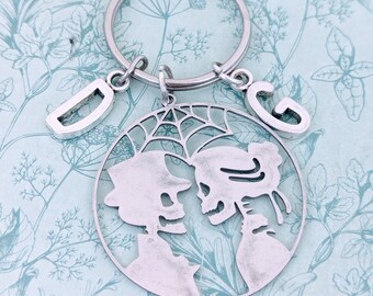 May include: A silver skeleton couple keychain with a spiderweb design. The keychain has two charms with the letters "D" and "G" hanging from it.
