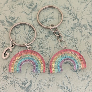 Peut inclure: Deux porte-clés en forme d'arc-en-ciel avec des anneaux argentés. Un porte-clés a un charm en argent en forme de lettre "G". Les breloques arc-en-ciel présentent un design coloré et translucide avec des teintes roses, jaunes, vertes et bleues.