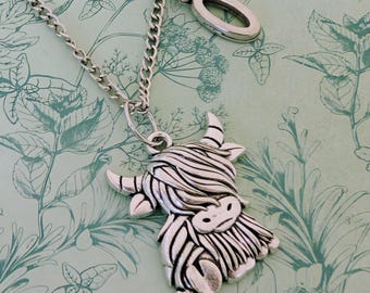 May include: A silver-toned necklace featuring a Highland cow pendant. The pendant is intricately detailed with striped fur and horns. The necklace has a chain with a lobster clasp and a decorative oval charm. The background has a light green floral pattern.